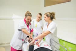 Ornella infirmière montre à deux étudiantes en soins infirmiers comment bander en épi les jambes de Julien
